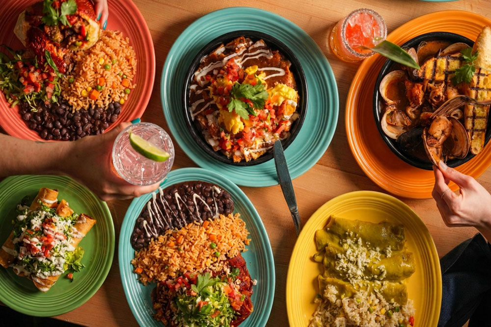 Overhead view of colorful Mexican dishes including tacos, enchiladas, rice, black beans, grilled seafood, and cocktails.