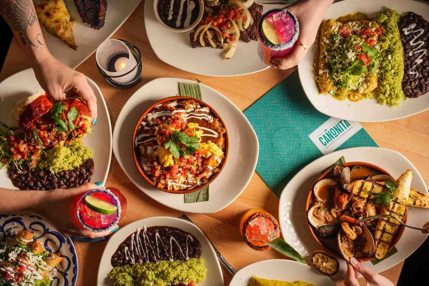 Overhead view of a table filled with colorful Mexican dishes, seafood, rice, beans, and bright cocktails.