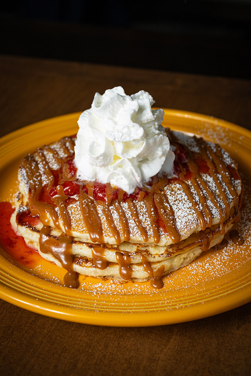 Stack of pancakes topped with strawberry sauce, caramel drizzle, powdered sugar, and whipped cream.