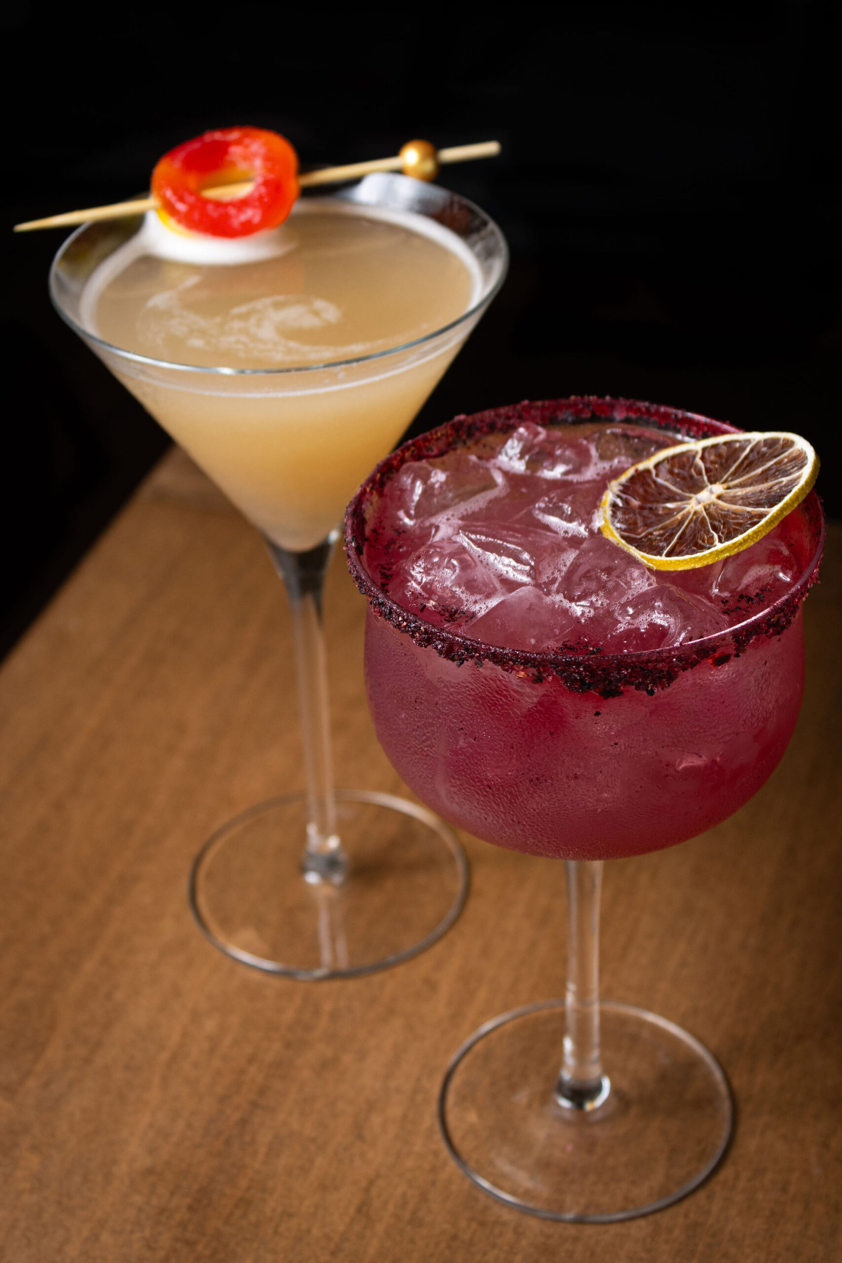 Two colorful cocktails, one pink with a citrus garnish and one pale cocktail garnished with a red pepper slice.